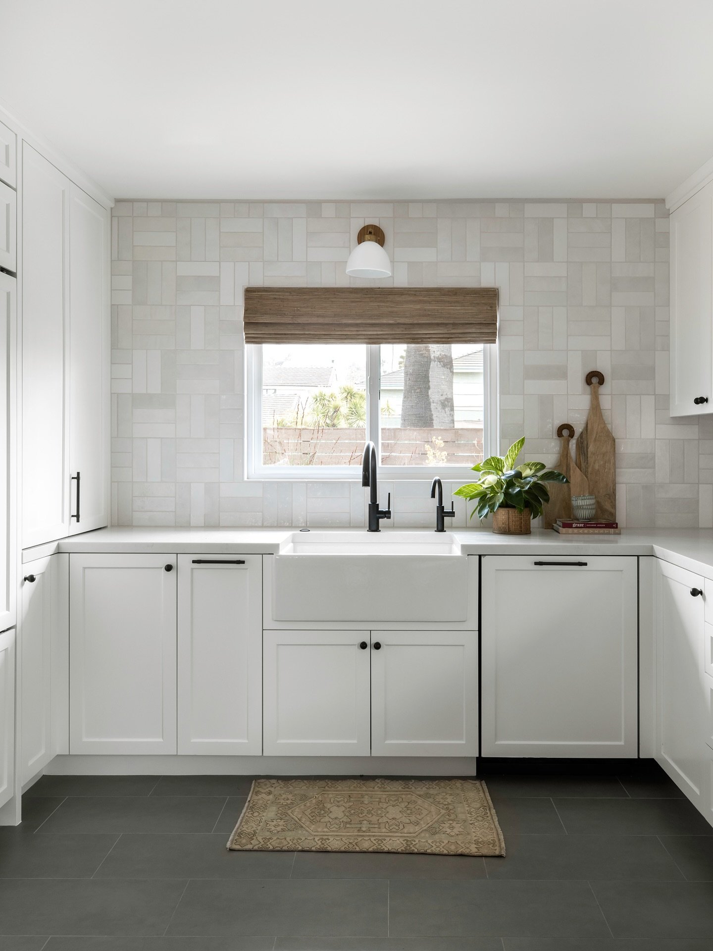 White kitchen featuring a rectangular white tile backsplash in a basketweave pattern, white shaker cabinets with black hardware, a farmhouse sink, and dark gray flooring.