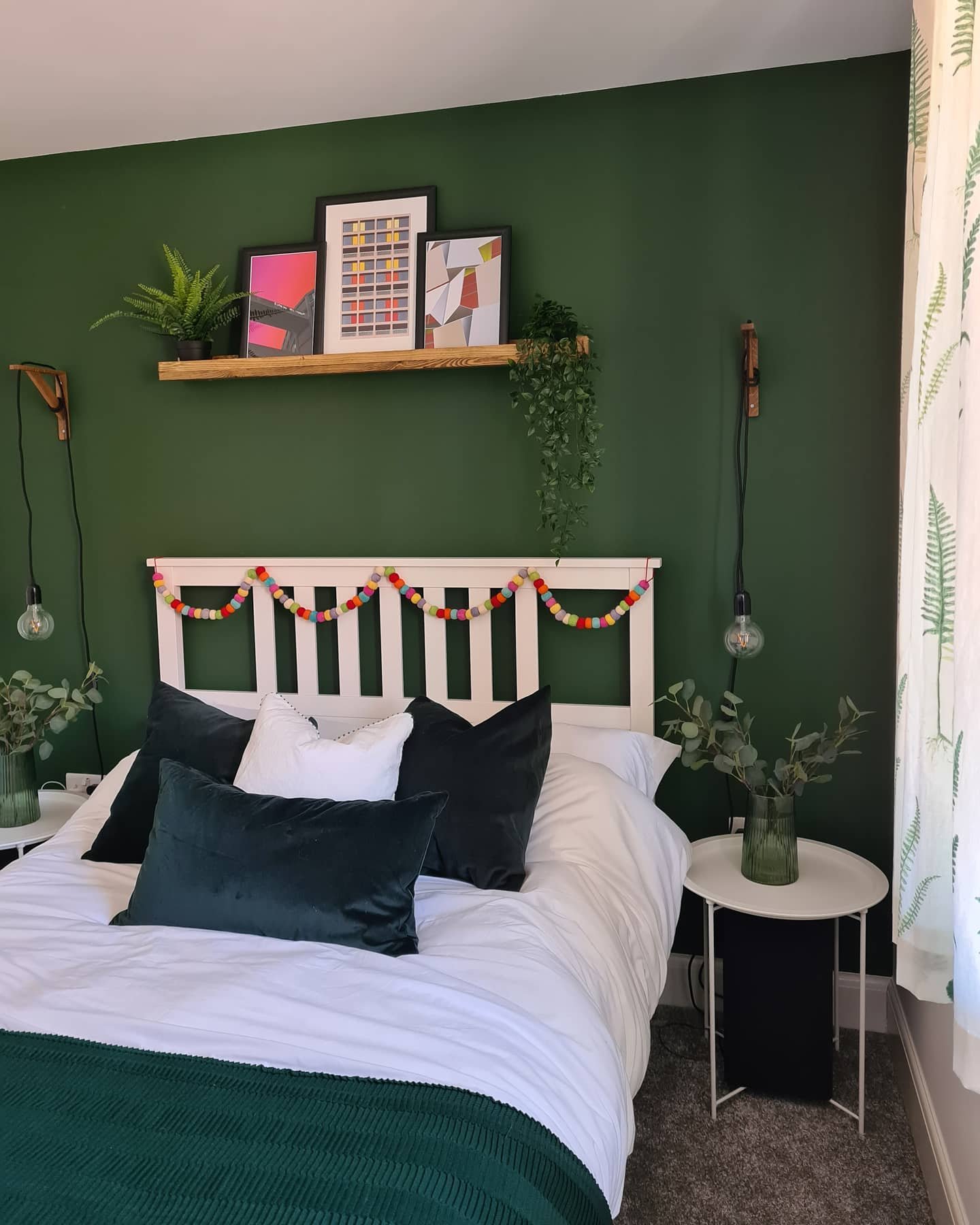 Bedroom featuring a solid dark emerald green wall, a white slatted headboard decorated with a colorful pom-pom garland, and a wooden floating shelf gallery.