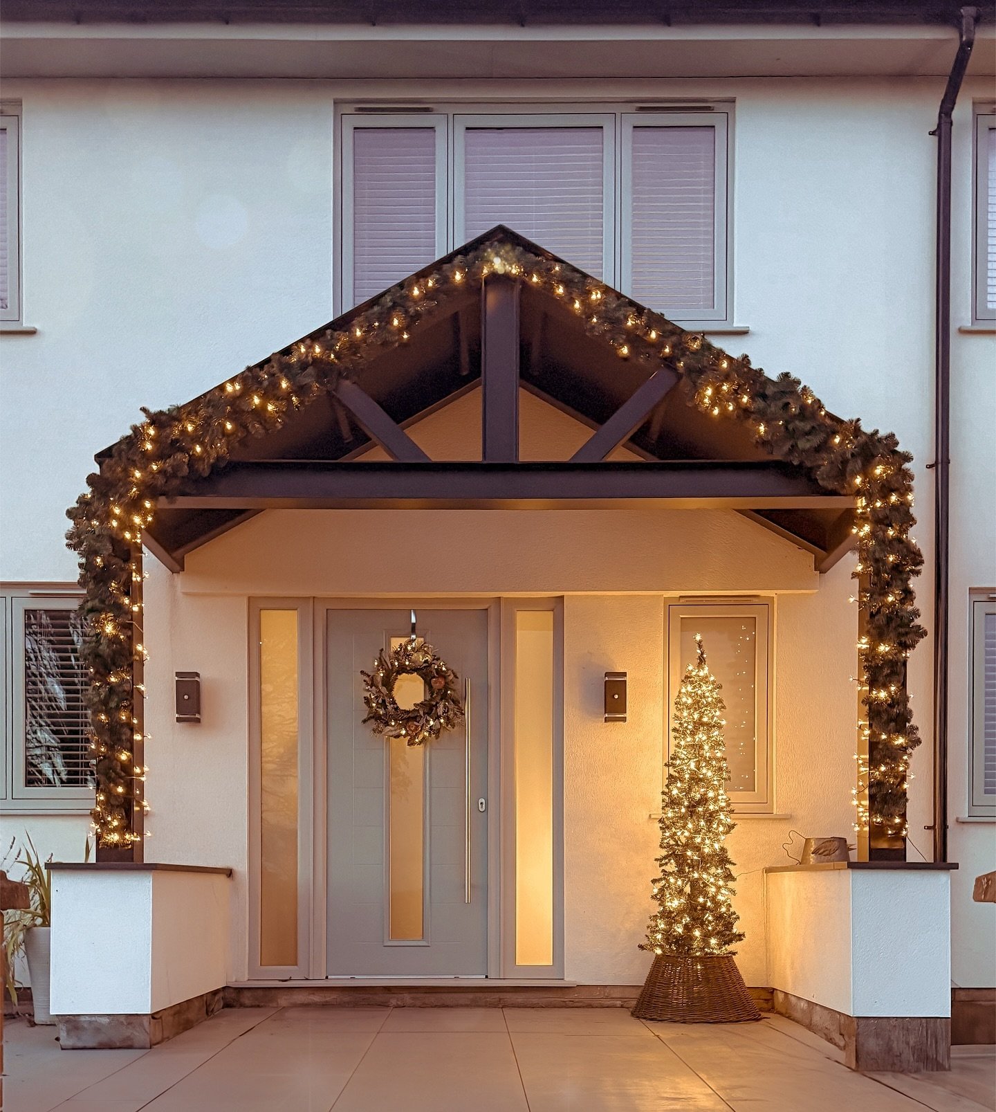 A covered entryway is framed by thick, pre-lit evergreen garland draped over the awning and down the supporting posts, with a tall, slim lighted Christmas tree standing by the light gray door.