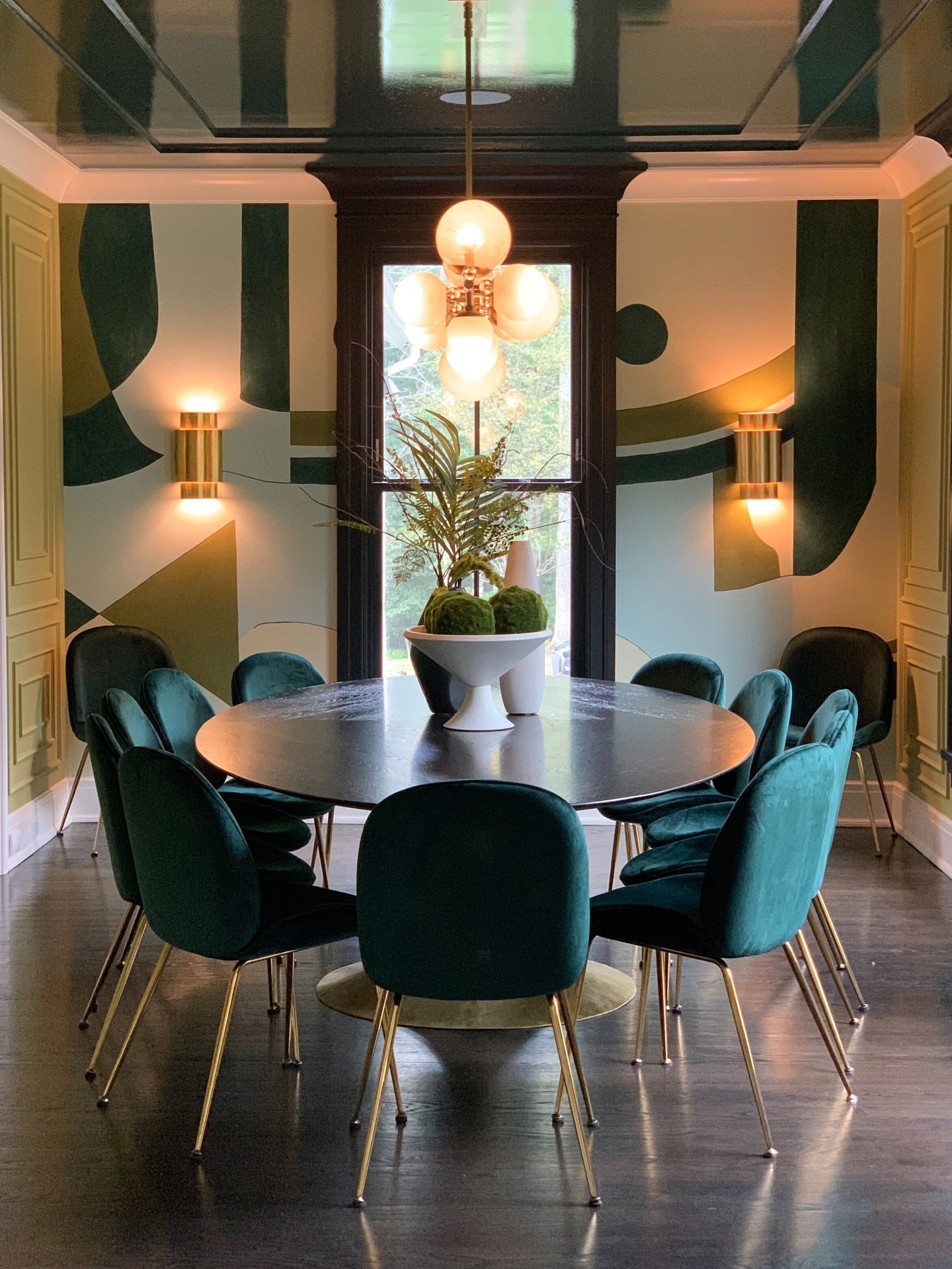 A luxurious dining room with a custom abstract mural in greens and tans, a dark oval table, and emerald green velvet chairs with gold legs, beneath a glossy black coffered ceiling.
