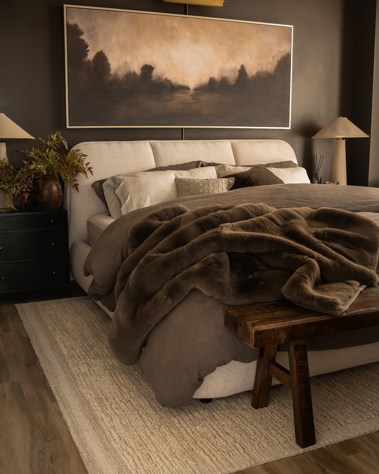 Dark bedroom with matte espresso brown walls, a chunky cream upholstered bed, dark brown linen bedding, a heavy faux fur throw, and an abstract landscape painting.