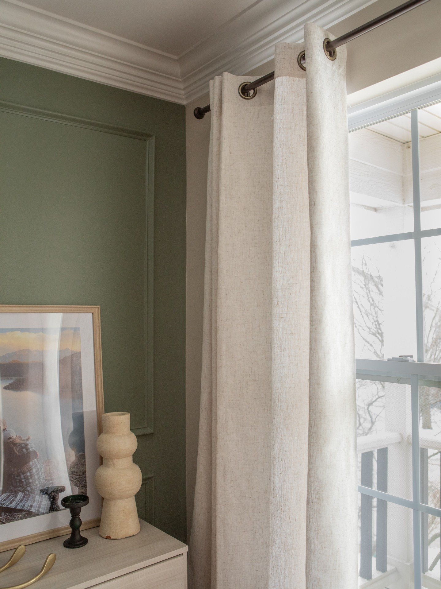 Close-up of a simple off-white linen texture curtain panel with grommet heading, contrasting against a dark sage green paneled wall.