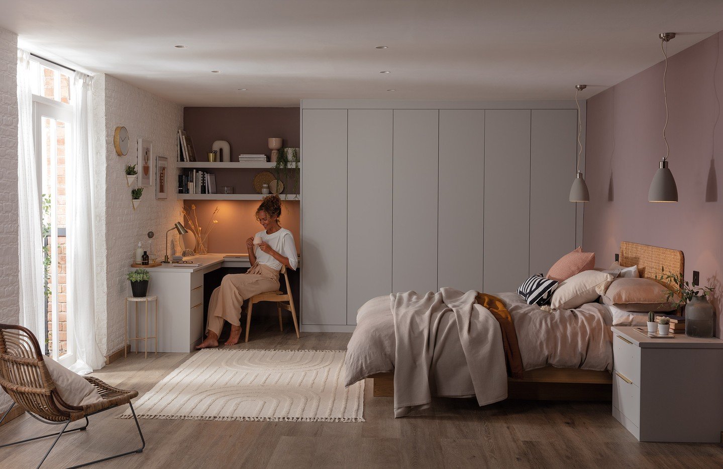 Wide shot of a modern bedroom with a large, seamless wall of light gray fitted wardrobes, contrasting with a small desk and open shelving nook.