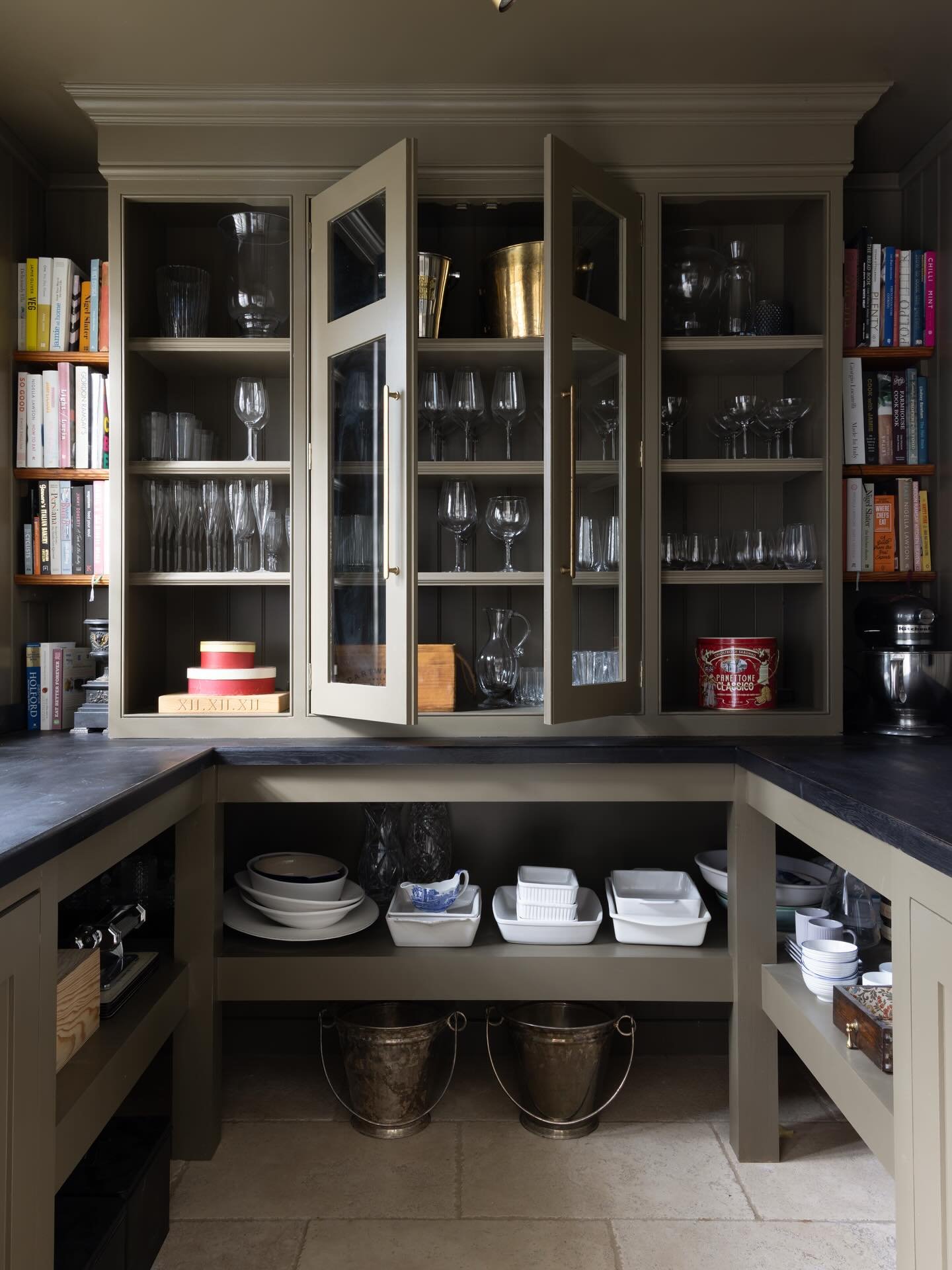 Dark, moody kitchen with open U-shaped counter and glass-front upper cabinets displaying rows of glassware, flanked by open cookbook shelving.
