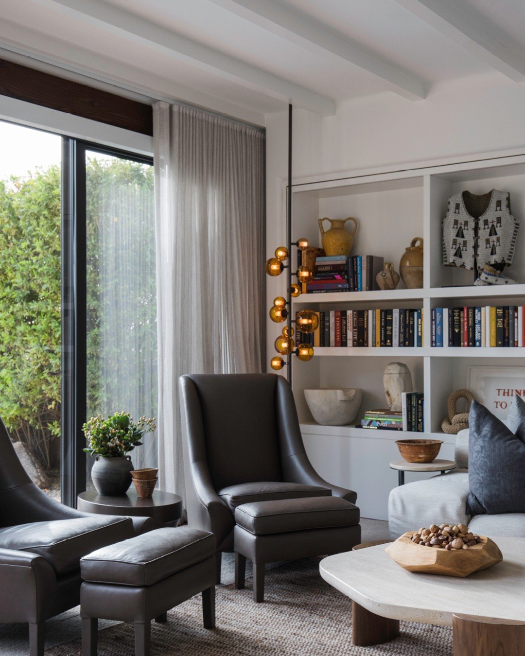 Luxury reading area with two dark brown leather high-back armchairs and matching ottomans, next to a large sliding glass door and white built-in bookshelves.