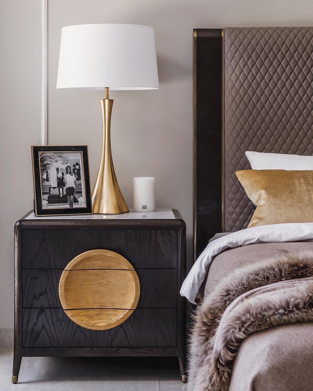 Dark black wood nightstand with a light wood circular inset accent on the top drawer, next to a diamond-quilted headboard.