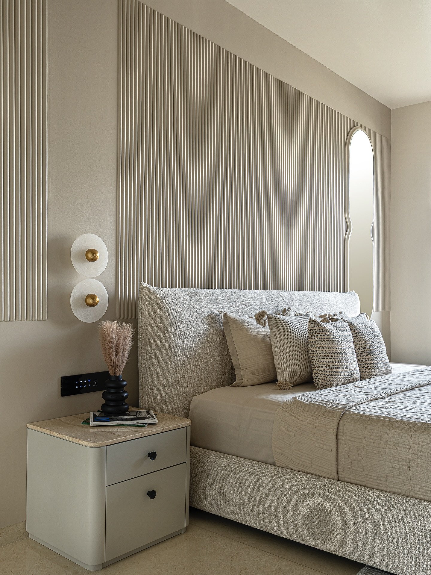 A contemporary beige bedroom featuring a massive architectural fluted wall panel behind the bed, a textured upholstered bed frame, and modern spherical wall sconces.