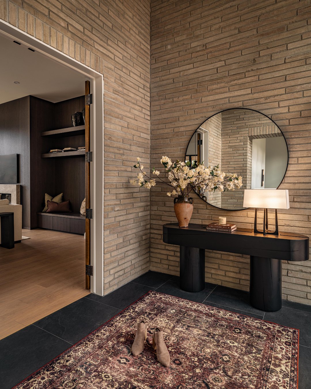 A hallway corner with exposed tan brick walls, a round black mirror, a black console table with cylindrical legs, and a vintage-style rug on a black tile floor.