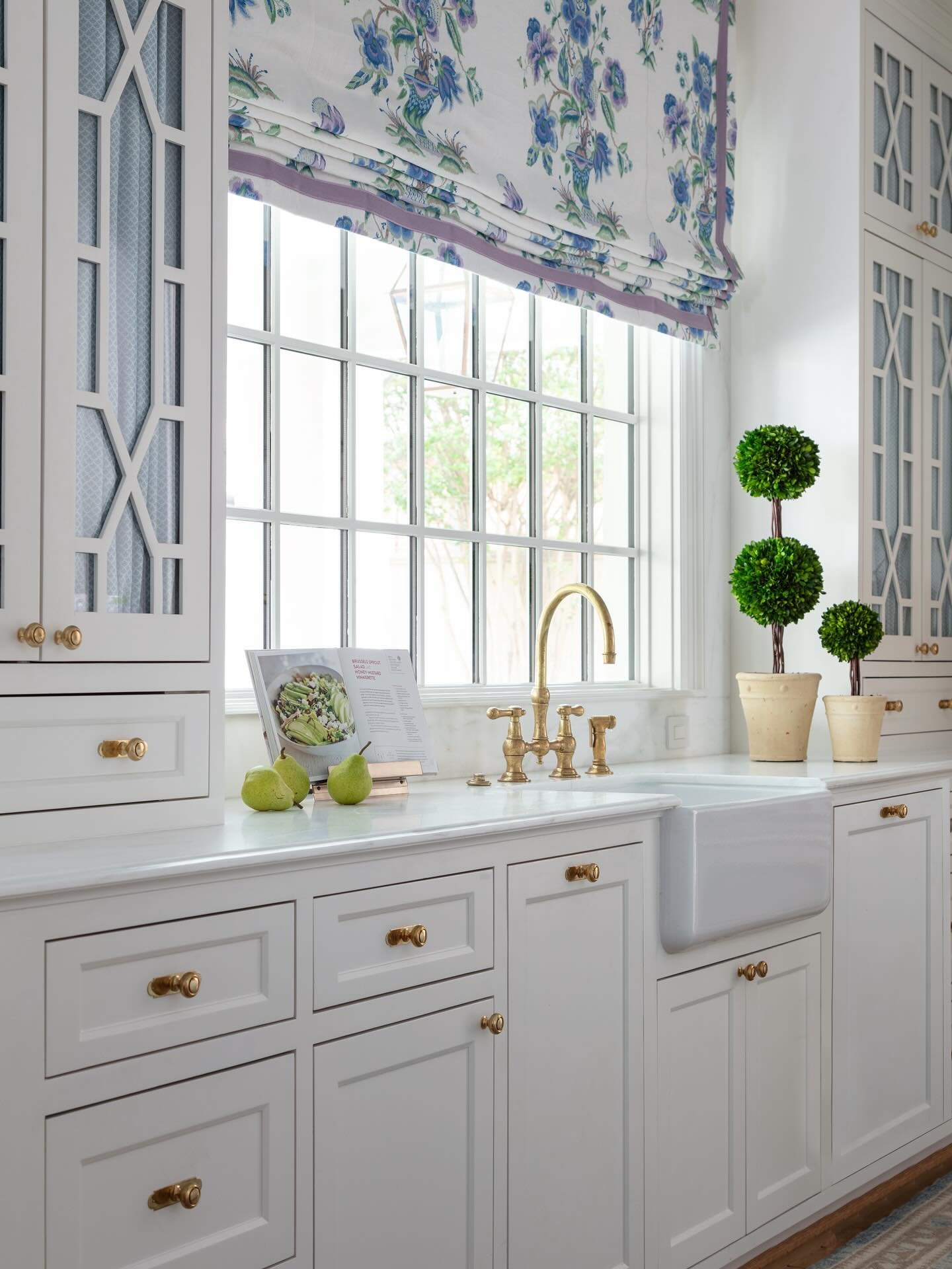 Elegant white kitchen with gold hardware and a farmhouse sink. A grid window is topped with a blue and purple floral patterned Roman shade, with small potted topiaries on the counter.