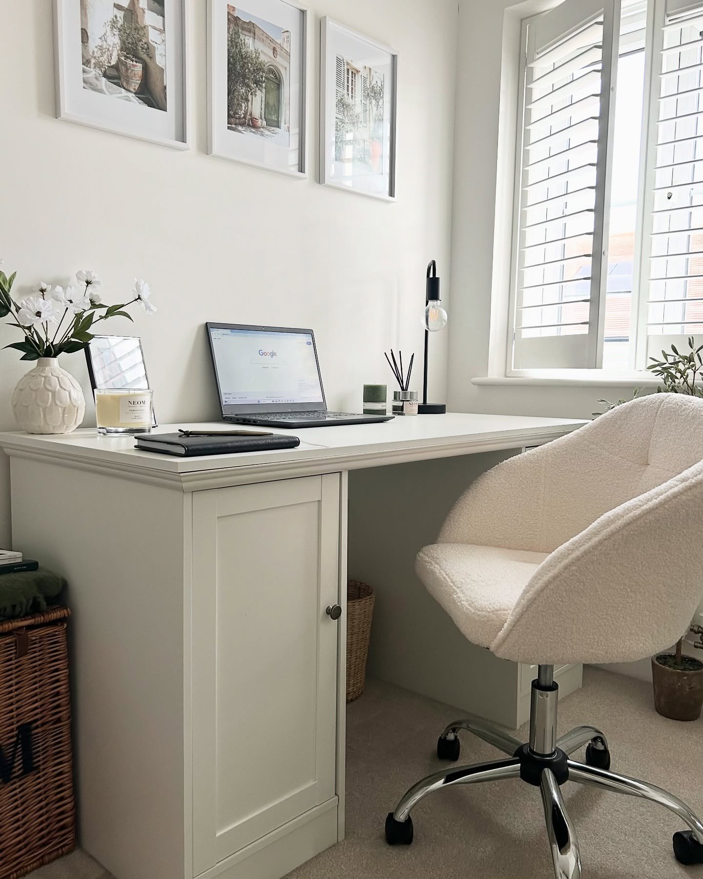 Bright home office with a white pedestal desk, a cream teddy-fabric rolling chair, white plantation shutters, and a trio of framed prints above the desk.