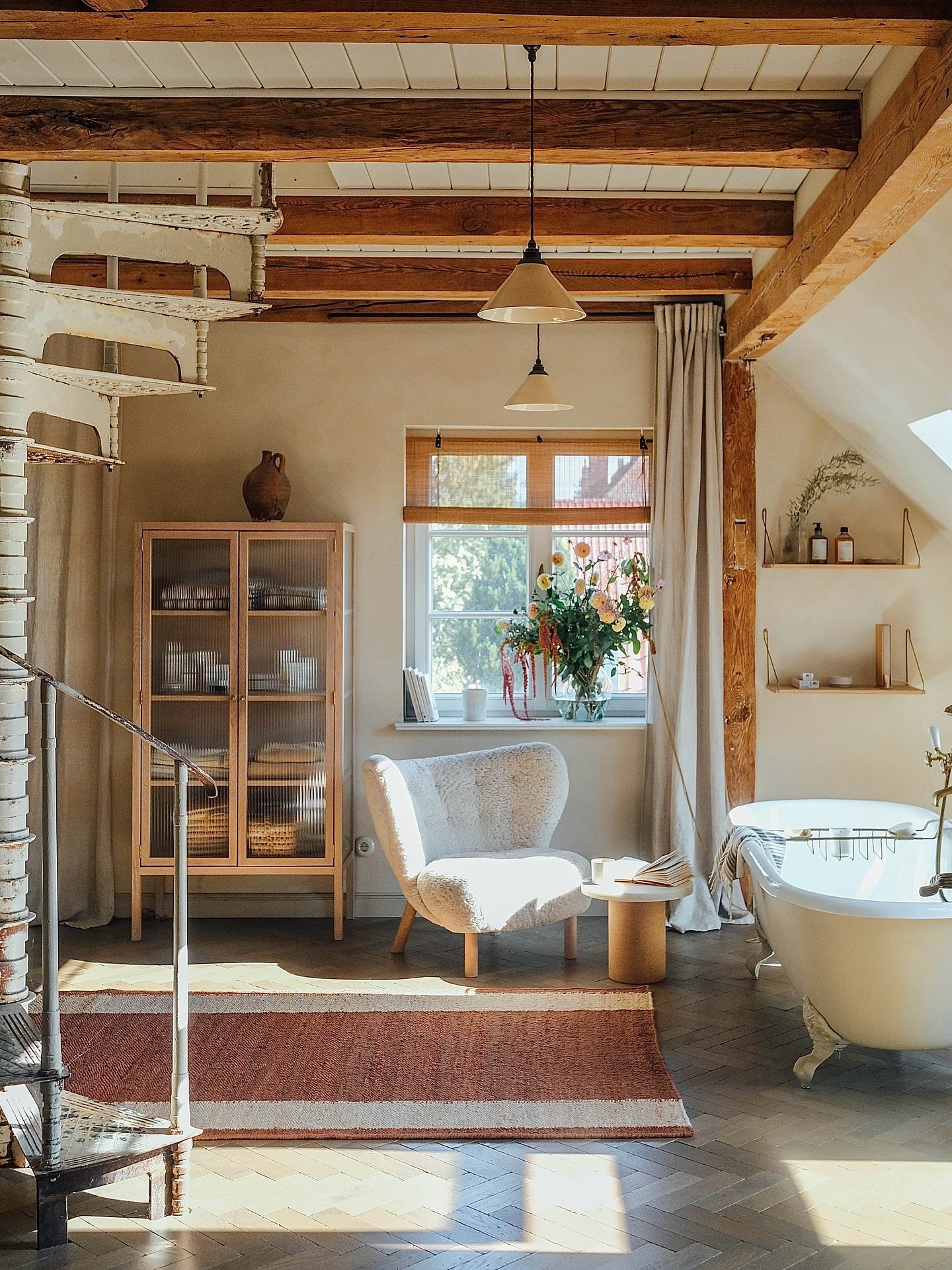 The room features striking exposed wood ceiling beams and a unique spiral staircase. A freestanding clawfoot tub sits under a sloped ceiling. A shaggy white accent chair creates a comfortable reading nook. The rug features a simple, yet striking striped pattern. The terracotta red and beige colors echo the warmth of the wood. This flat-weave rug adds a cozy, grounded feeling to the architectural space.