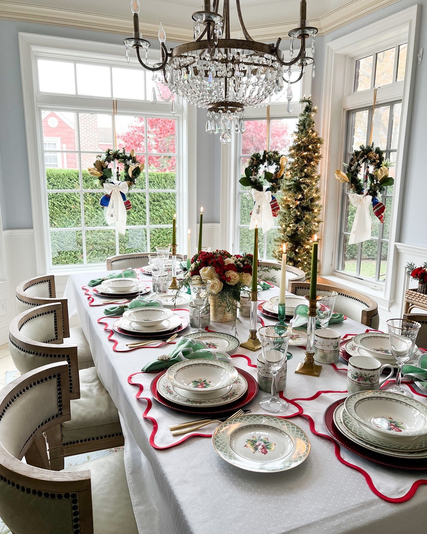 A bright dining table set with a white tablecloth, featuring red scalloped placemats, vintage floral and holly-patterned china, dark green napkins, and a centerpiece of tall olive-green taper candles in brass holders and a small bouquet of roses and holly.