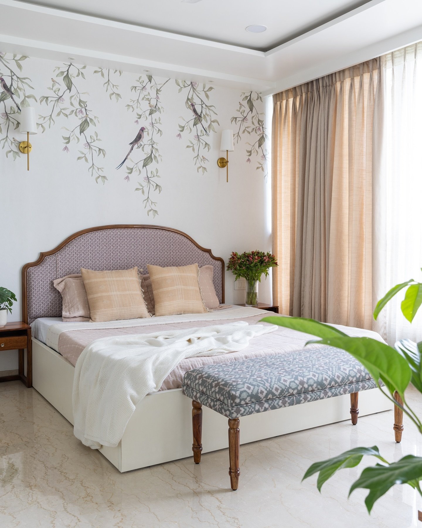 A bright bedroom with a bed featuring an arched pink headboard, a nature-themed mural on the wall, and a small, upholstered bench with a blue and gray ikat print at the foot of the bed.