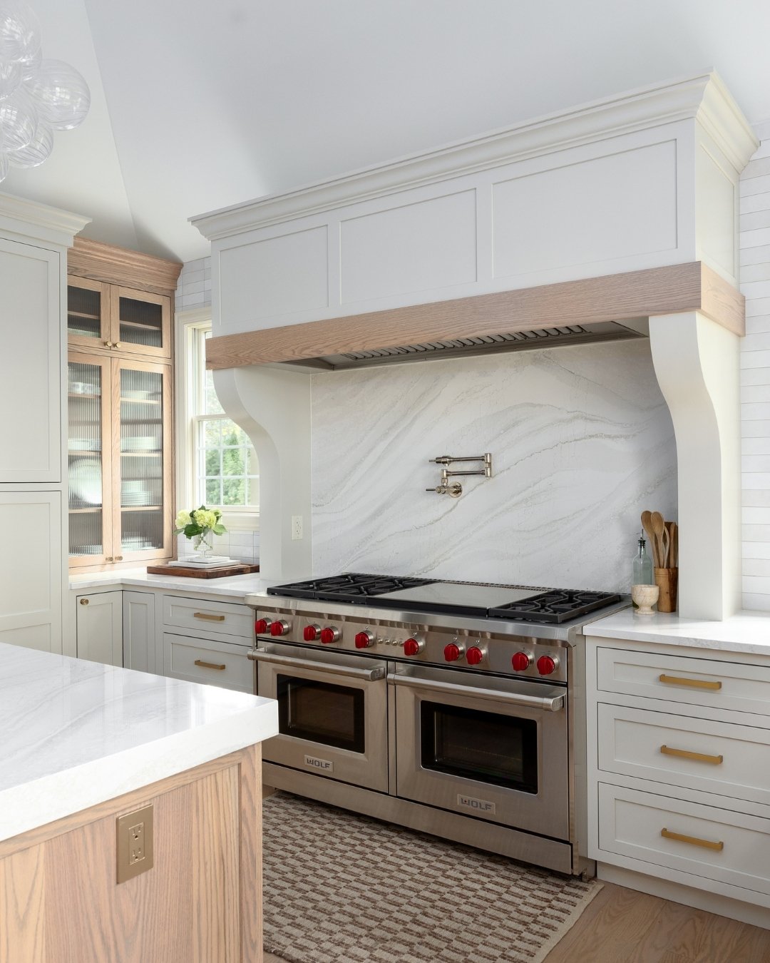 Luxury kitchen with a full-slab white quartz backsplash framed by white subway tile, a white and wood integrated range hood, a stainless steel Wolf range, and light cabinetry with gold hardware.
