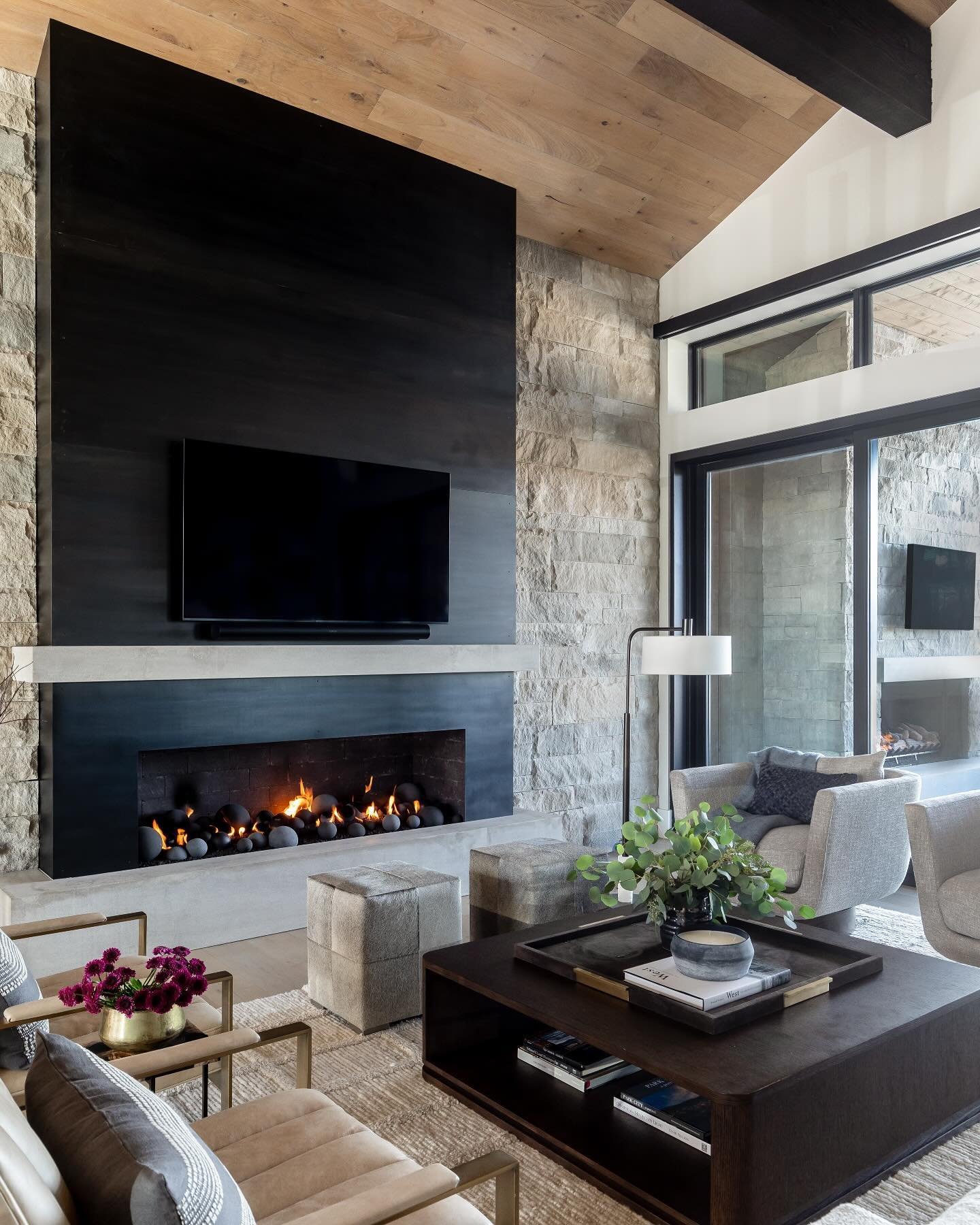 Large contemporary living room with a linear fireplace set into a floor-to-ceiling stone wall and dark metal paneling, with vaulted wood ceilings.