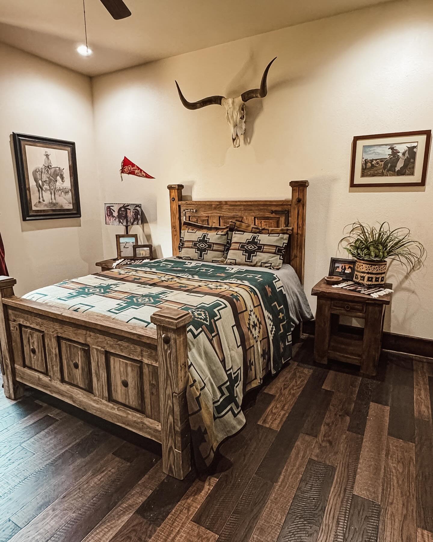 A Western bedroom featuring a longhorn skull mounted over a sturdy, light-toned wood platform bed, Southwestern bedding, and framed cowboy art.