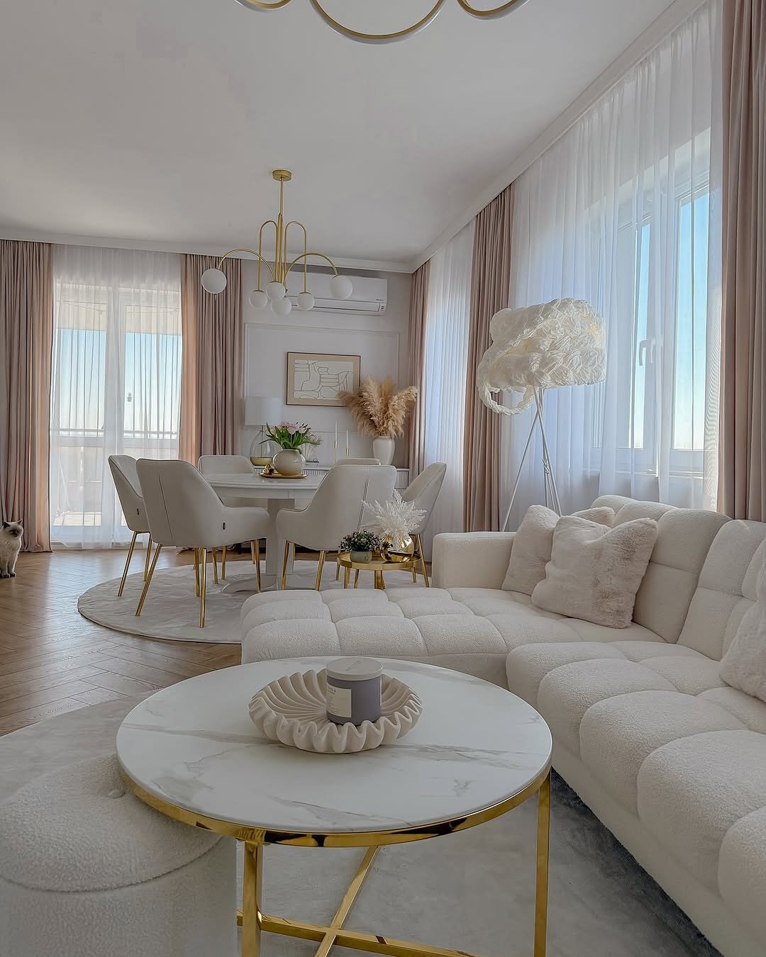 A luxurious open-concept living and dining area featuring a tufted white sectional, a round marble-top coffee table with a gold base, and matching dining furniture.