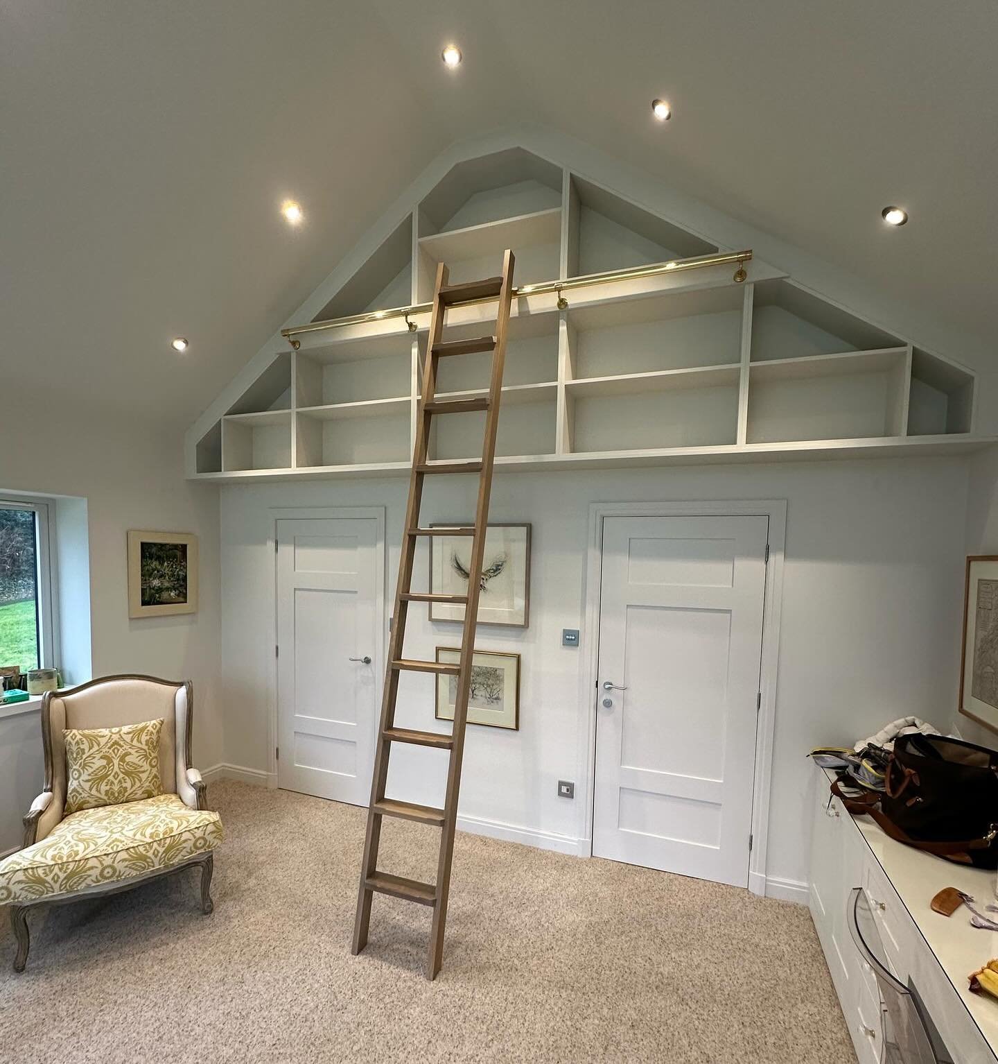Custom white built-in bookshelves following the inverted V-shape of a vaulted ceiling, situated high above two white doors, accessible by a tall wooden library ladder with a brass sliding rail.