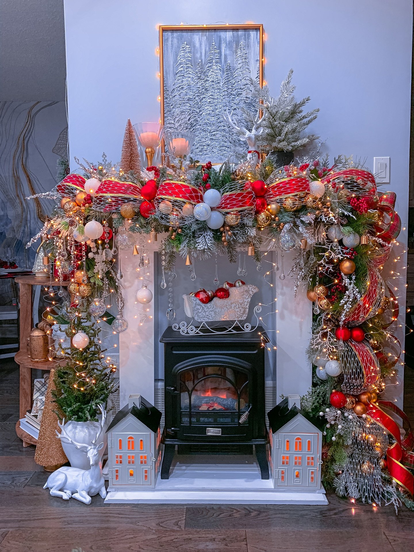 White fireplace with a black wood stove insert, surrounded by a heavy, frosted evergreen garland, wide red ribbon, gold ornaments, and two illuminated white house lanterns.