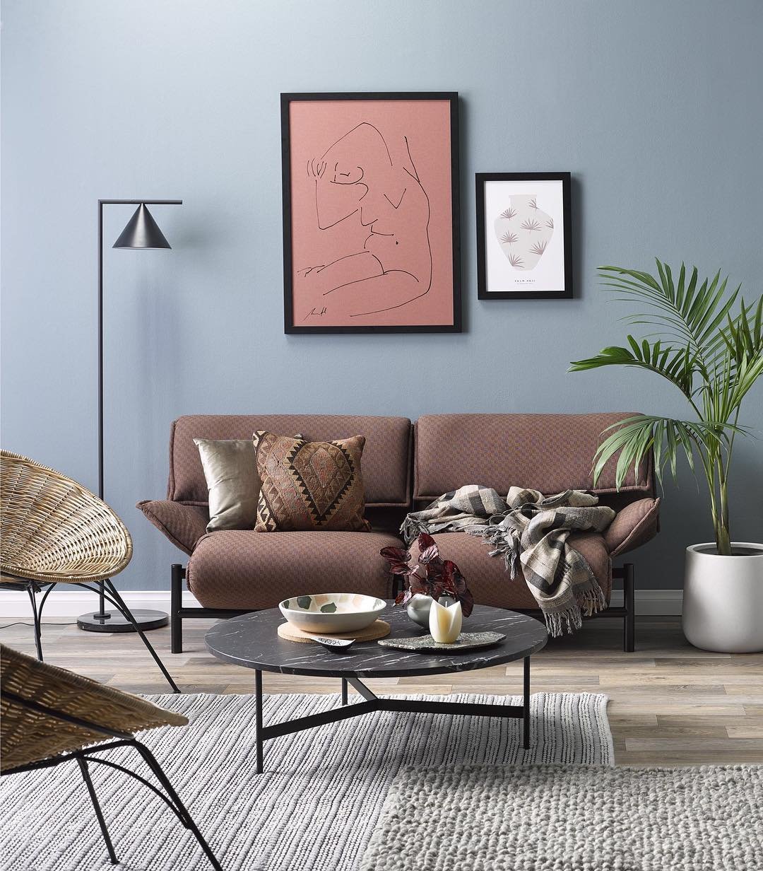 Small living area with a brown mid-century modern sofa, black marble coffee table, wicker chair, and pink abstract line art on a blue wall.