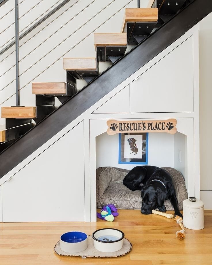A black dog resting in a custom-built, white sleeping den under modern open-tread stairs.