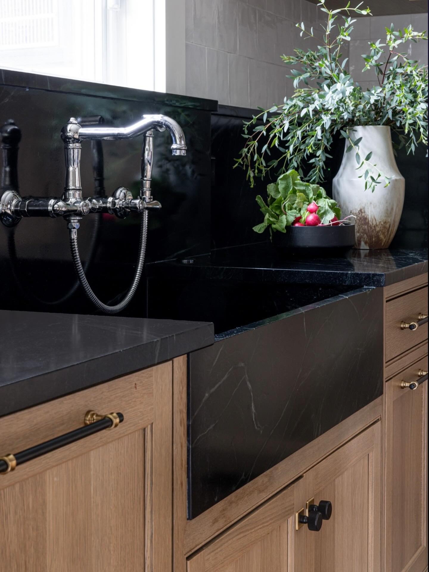 A luxurious single-bowl farmhouse sink carved from dark black stone with white veining, featuring a matching dark stone countertop and backsplash and a traditional wall-mounted chrome bridge faucet.