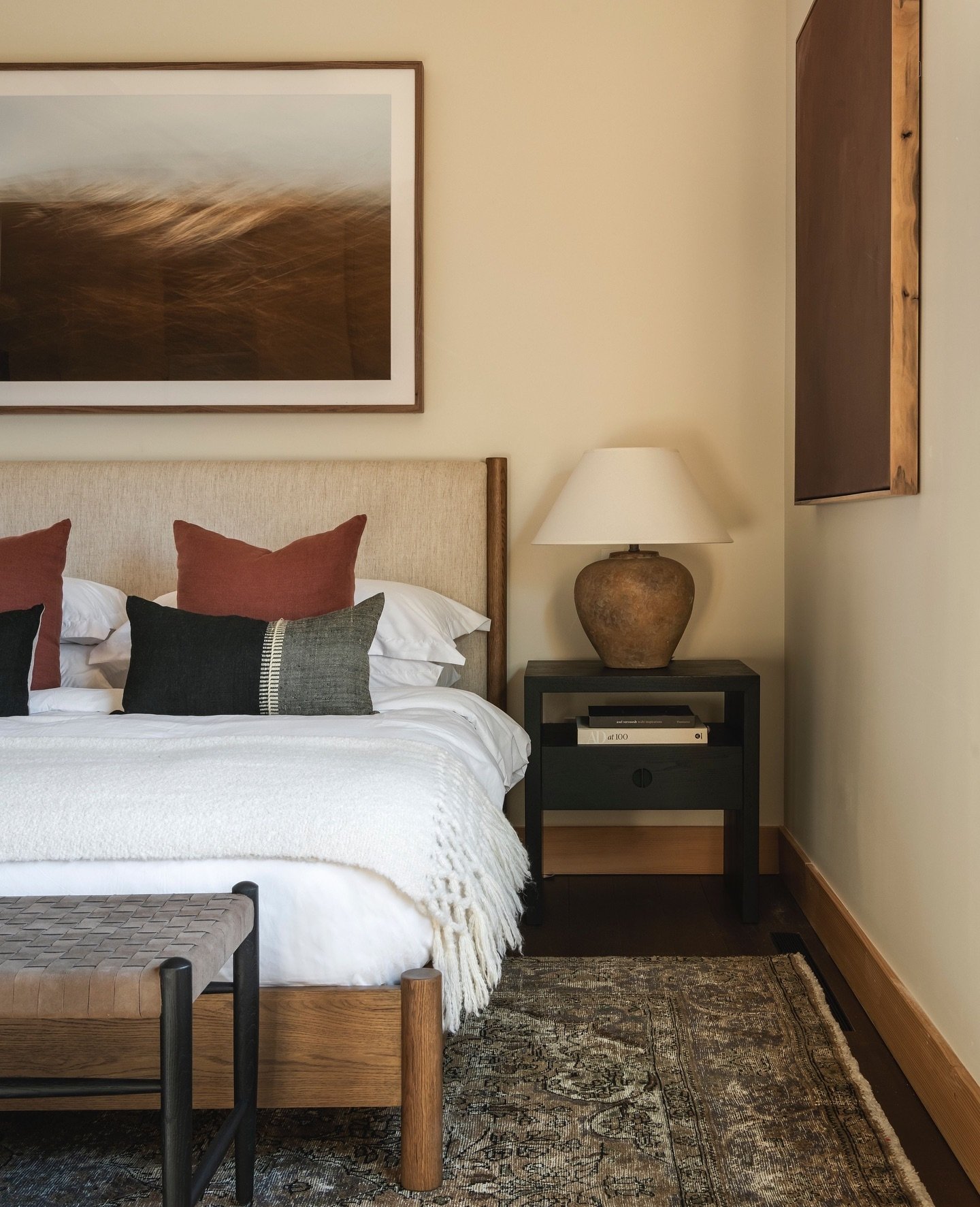 Simple black nightstand with one drawer and an open shelf, next to a neutral bed with earthy-toned pillows and a textured rug.