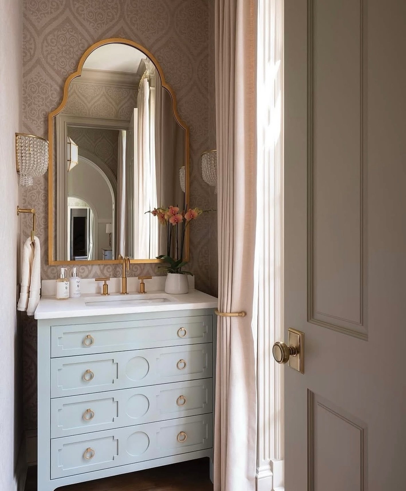 Light blue-gray vanity with gold ring pulls, set against subtle brocade-patterned wallpaper, with a large arched gold mirror.