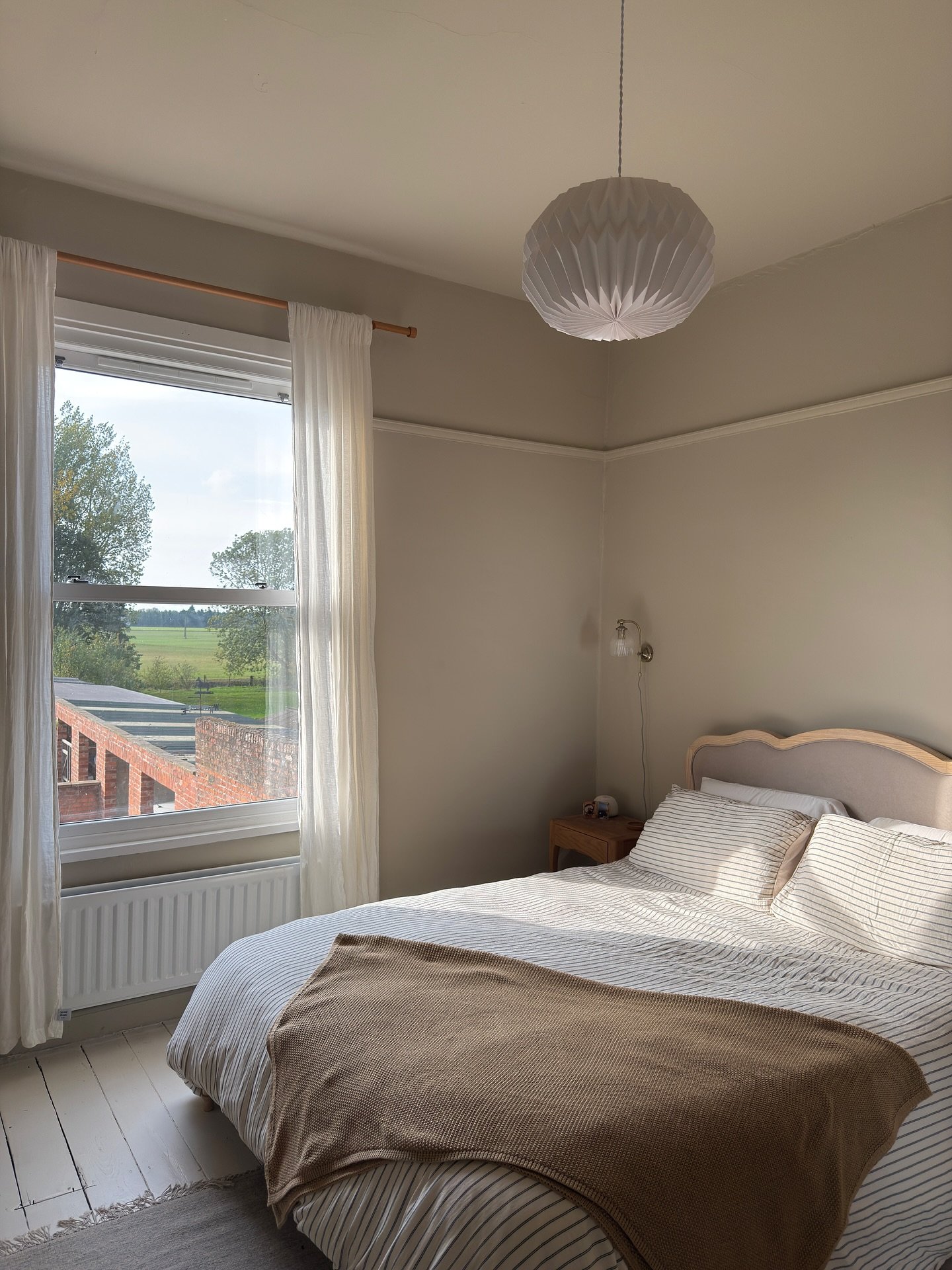 A simple, light-filled beige bedroom with a small wooden headboard, thin striped bedding, a dark brown throw blanket, a white paper lantern pendant, and a large window overlooking a brick building and green fields.