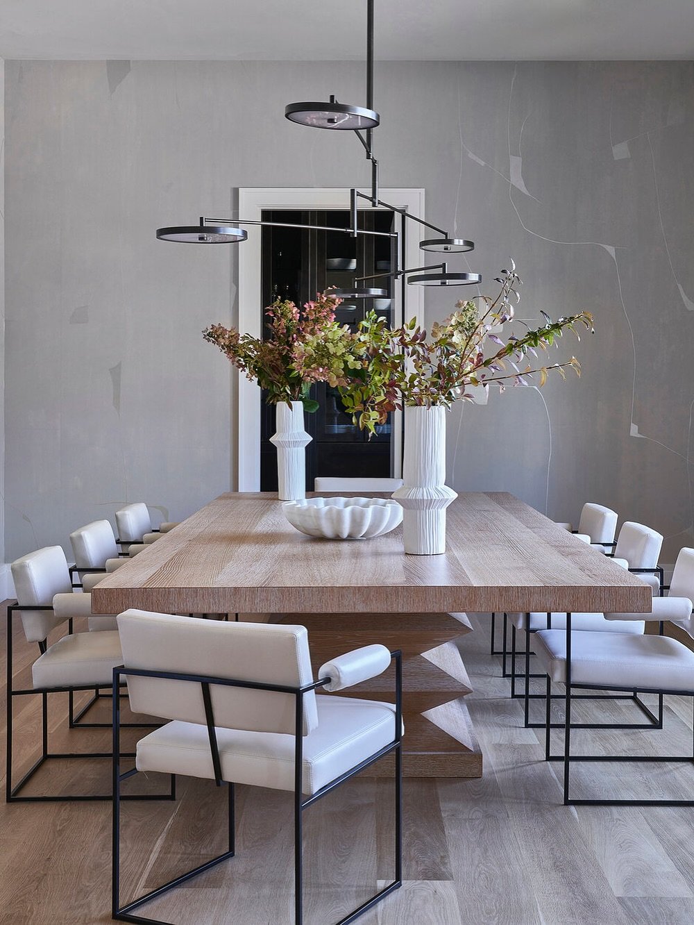 A minimalist dining room with a long light wood table on a sculptural base, sleek white leather chairs with black metal frames, and a modern, angular black chandelier.