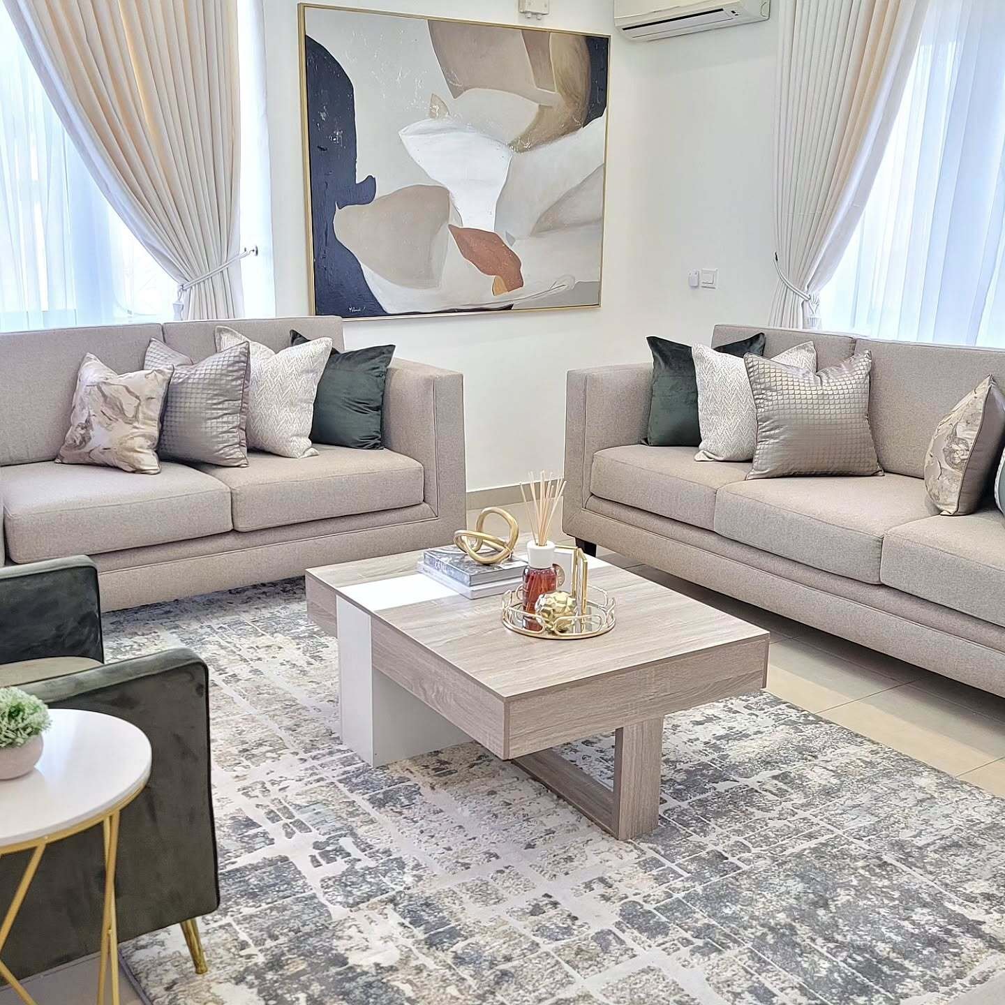 A symmetrical beige living room featuring two identical sofas, deep green velvet accent chairs, a modern gray wood coffee table, and large abstract artwork.