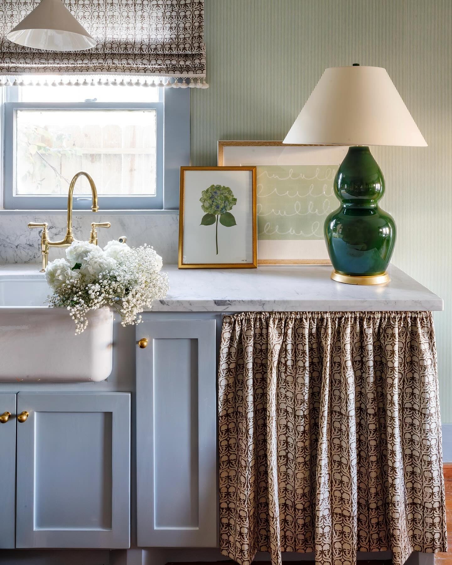 Kitchen sink area with pale blue cabinets, featuring a coordinated brown and cream patterned Roman shade on the window and a matching gathered fabric skirt under the counter.