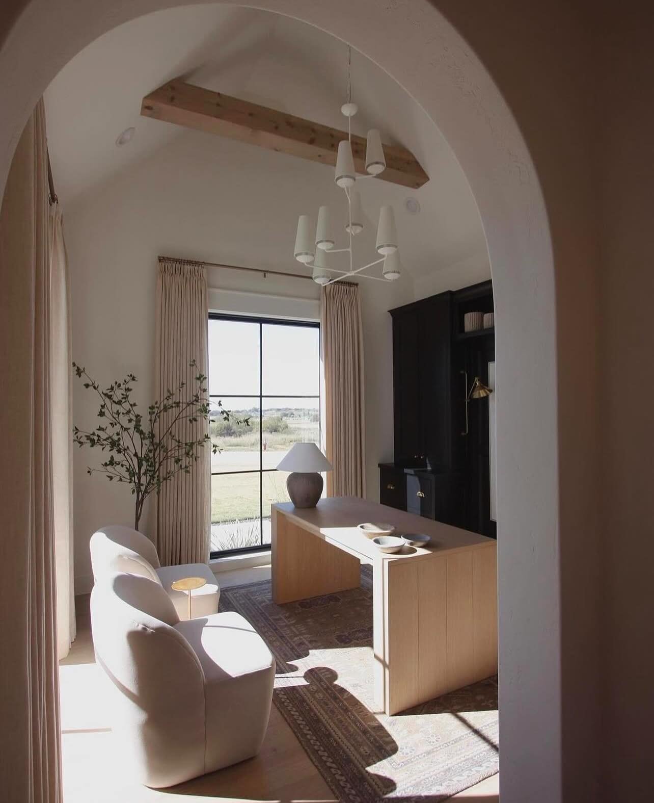 Home office with a vaulted ceiling, a light wood waterfall desk, a black built-in cabinet, a light cream upholstered armchair, and a large black-framed window.