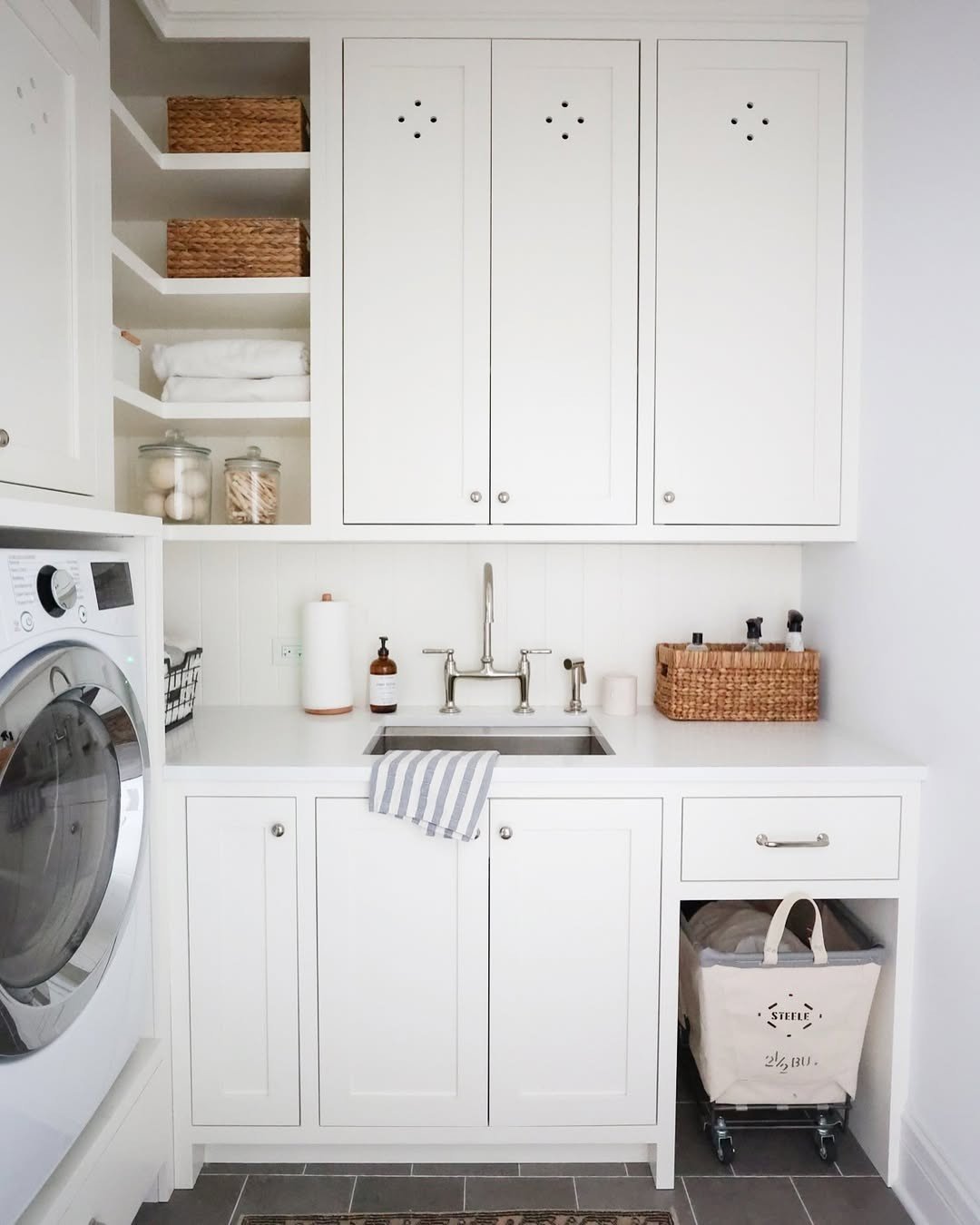 A small, crisp white laundry room featuring a stainless steel sink, Shaker-style cabinets, open shelving on the left, and a dedicated pull-out niche for a rolling laundry cart on the right.