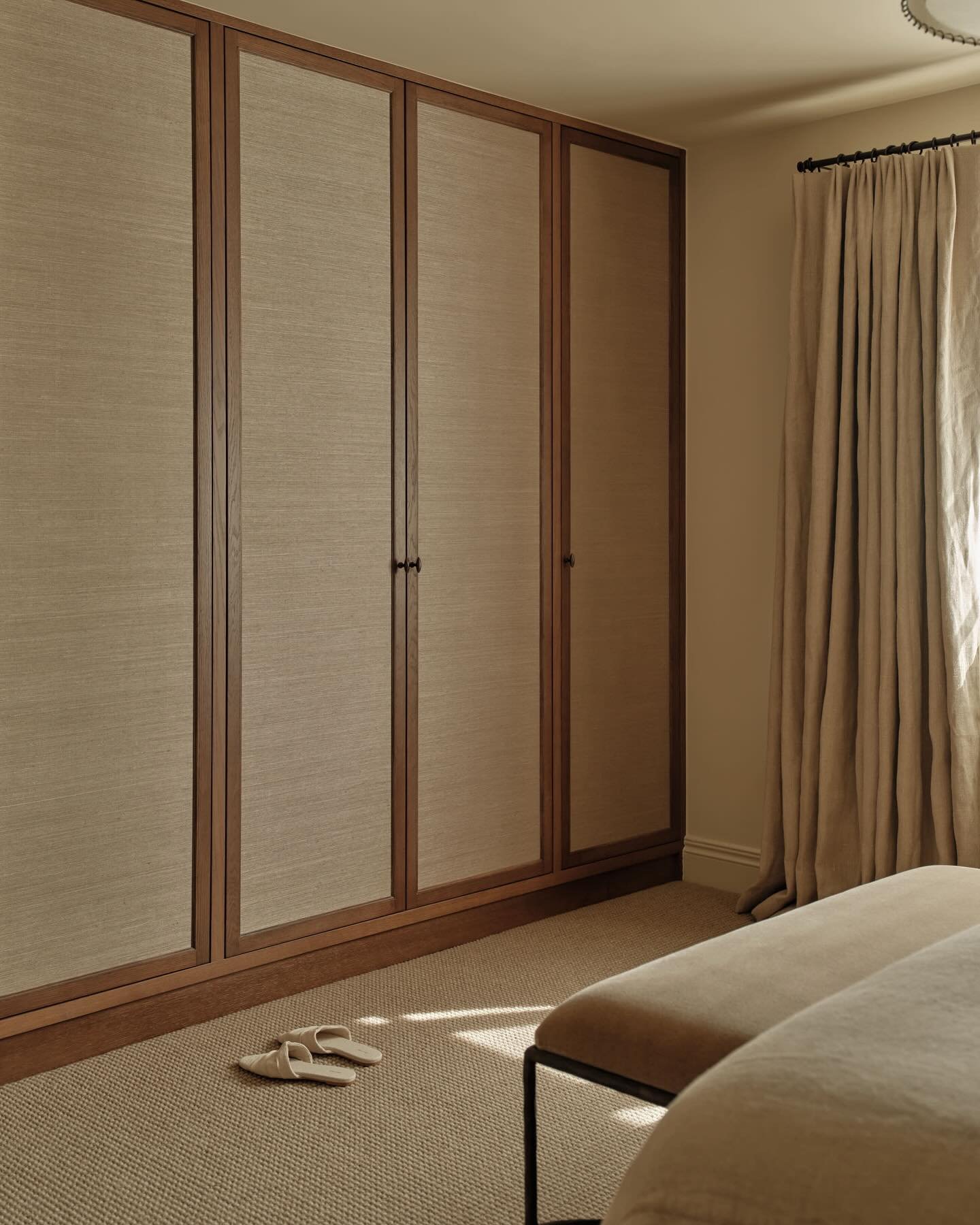 Full wall of built-in wardrobes with dark wood frames and inset panels covered in a neutral, woven textured fabric, creating a sophisticated wall feature.