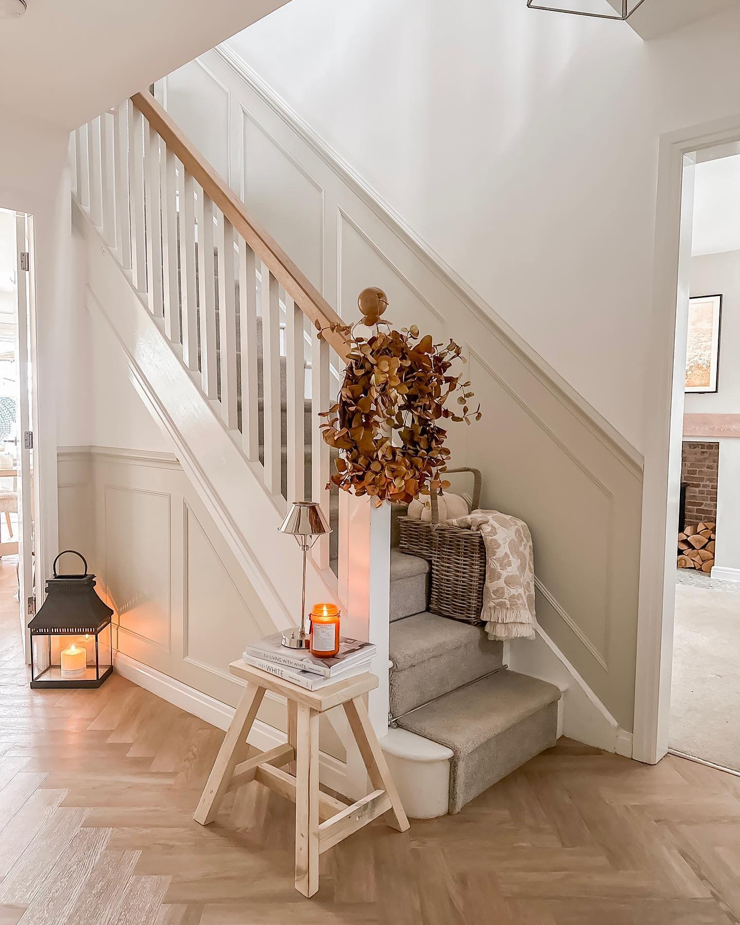 A staircase with light grey wainscoting and a carpet runner, decorated with a wooden stool holding books and a candle, a large black lantern, and a fall wreath hanging on the banister.