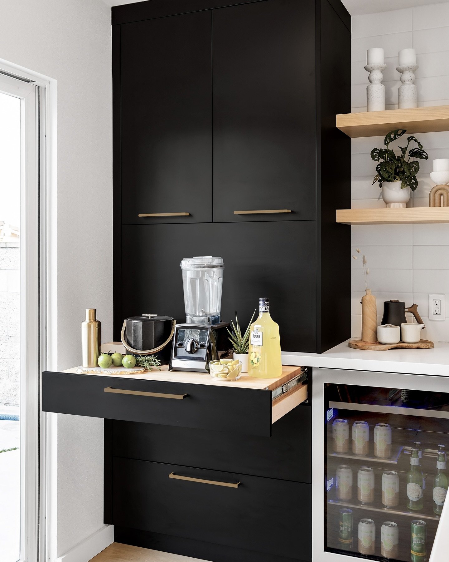 Modern black cabinetry with gold hardware, featuring a tall cupboard, floating shelves, and a horizontal pull-out drawer used as a bar prep surface.