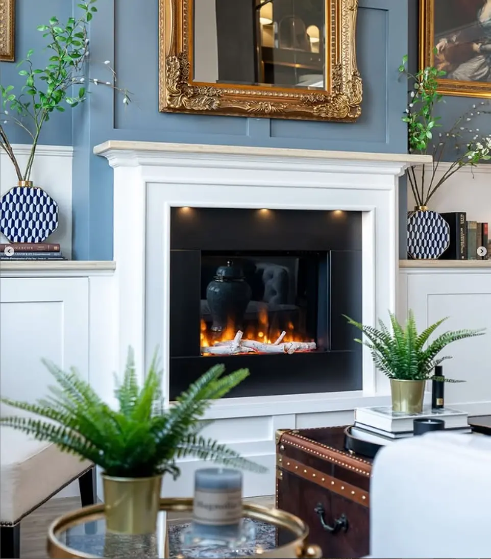 White wooden mantel surrounding an inset black electric fireplace displaying realistic glowing log flame effects in a room with blue paneled walls.
