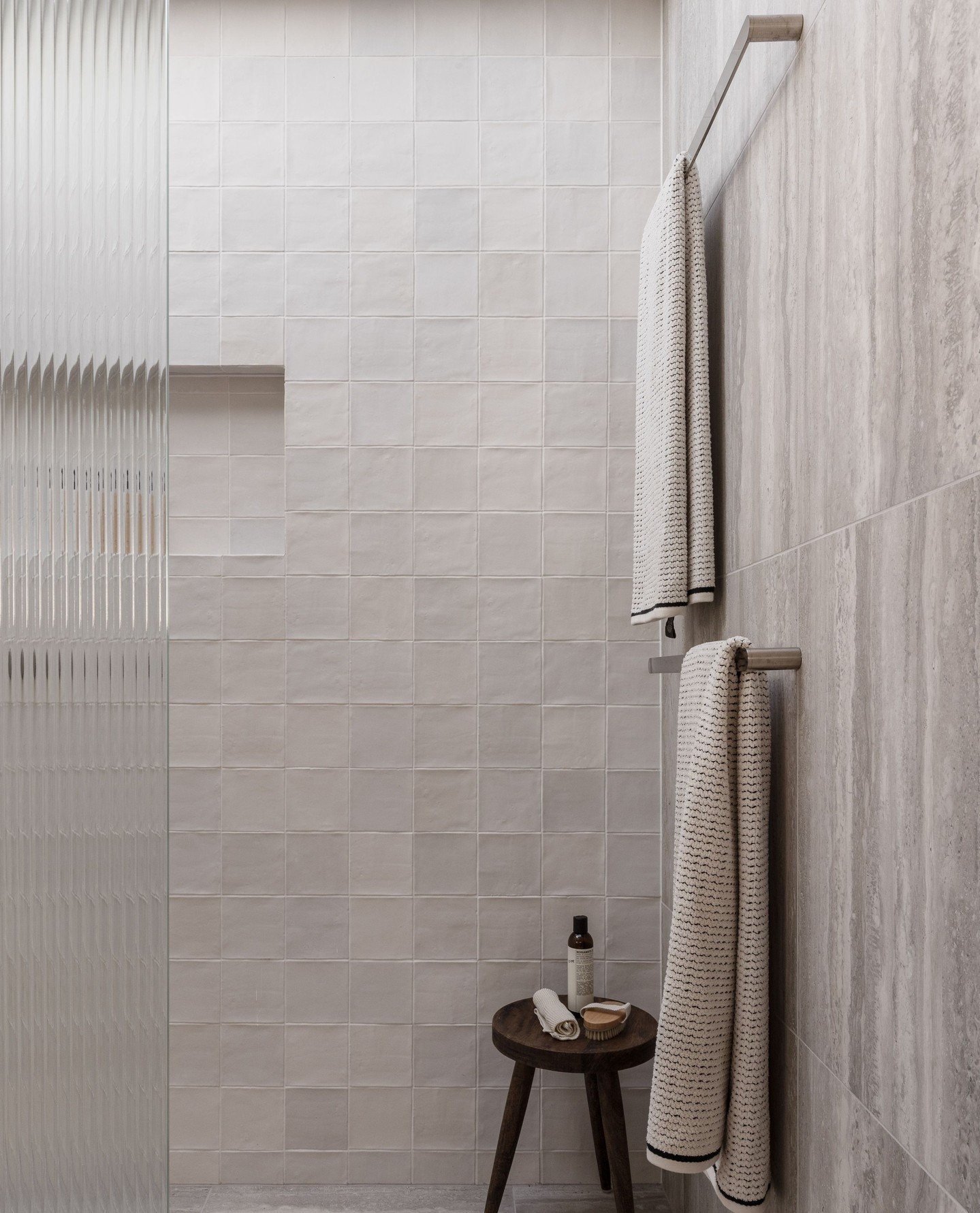 Two grey patterned towels hanging vertically on a sleek double stainless steel bar against a large format stone-look tile wall in a neutral bathroom.