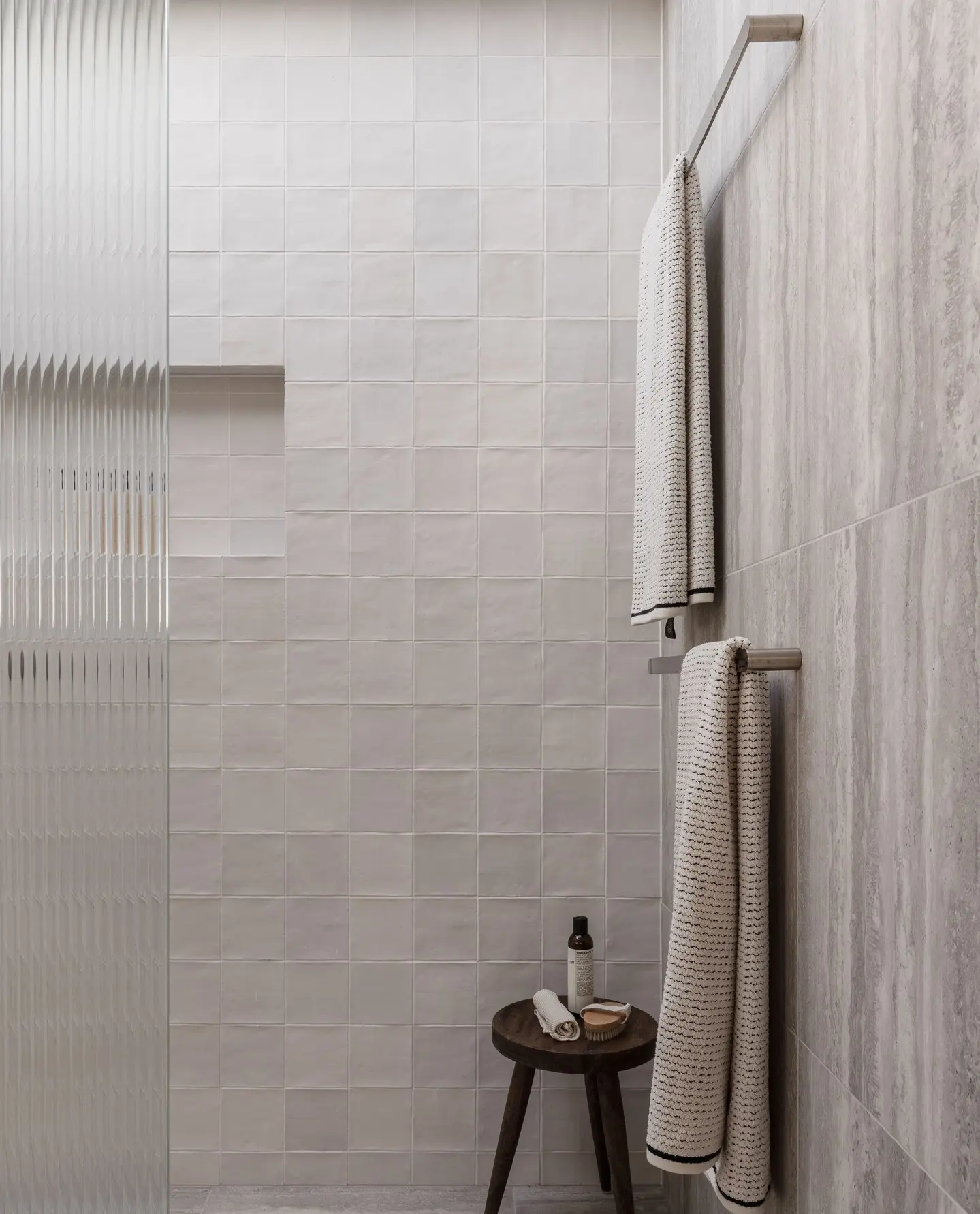 Two grey patterned towels hanging vertically on a sleek double stainless steel bar against a large format stone-look tile wall in a neutral bathroom.