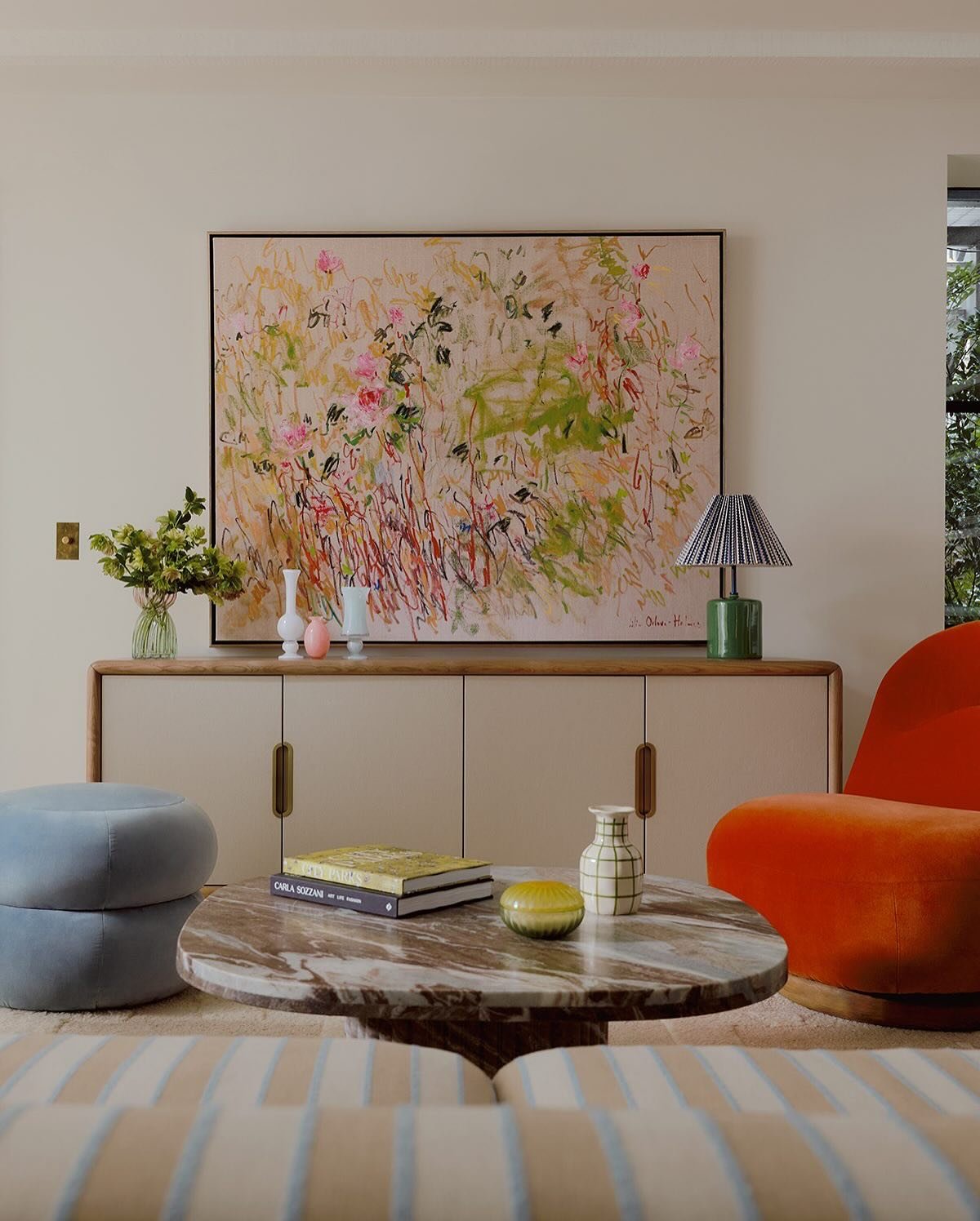 Horizontal framed impressionistic painting with pink and green brushstrokes, hung above a light beige media console next to an orange armchair and blue ottomans.