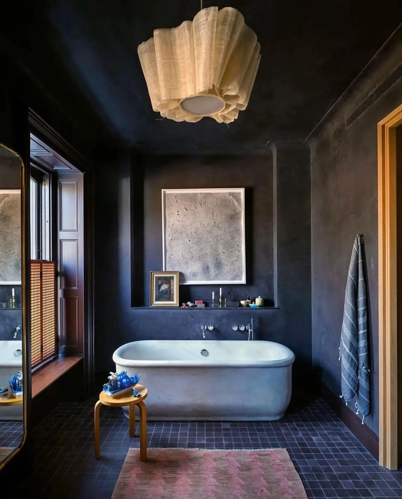 A dark, moody bathroom with matte black walls, a white soaking tub, and layered artwork on a recessed shelf.