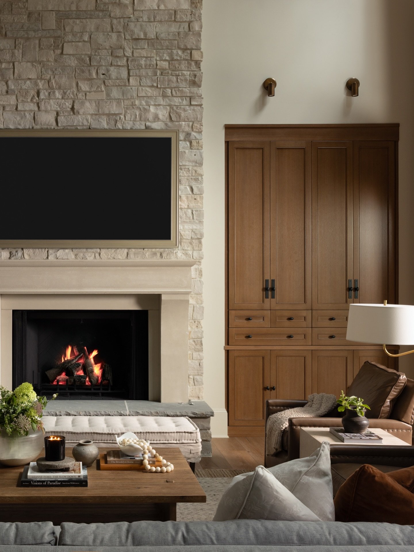 Living room with a full-height stacked stone fireplace, a large TV mounted above, custom light wood built-in cabinetry, and a leather armchair.