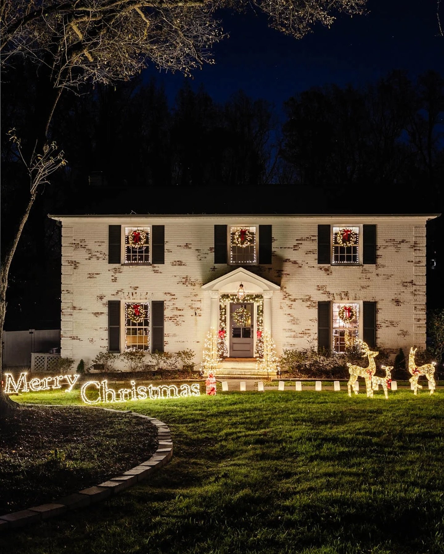 A white brick house decorated with wreaths and lights is fronted by a large, glowing, warm white "Merry Christmas" wire sign on the lawn, accompanied by two light-up reindeer.