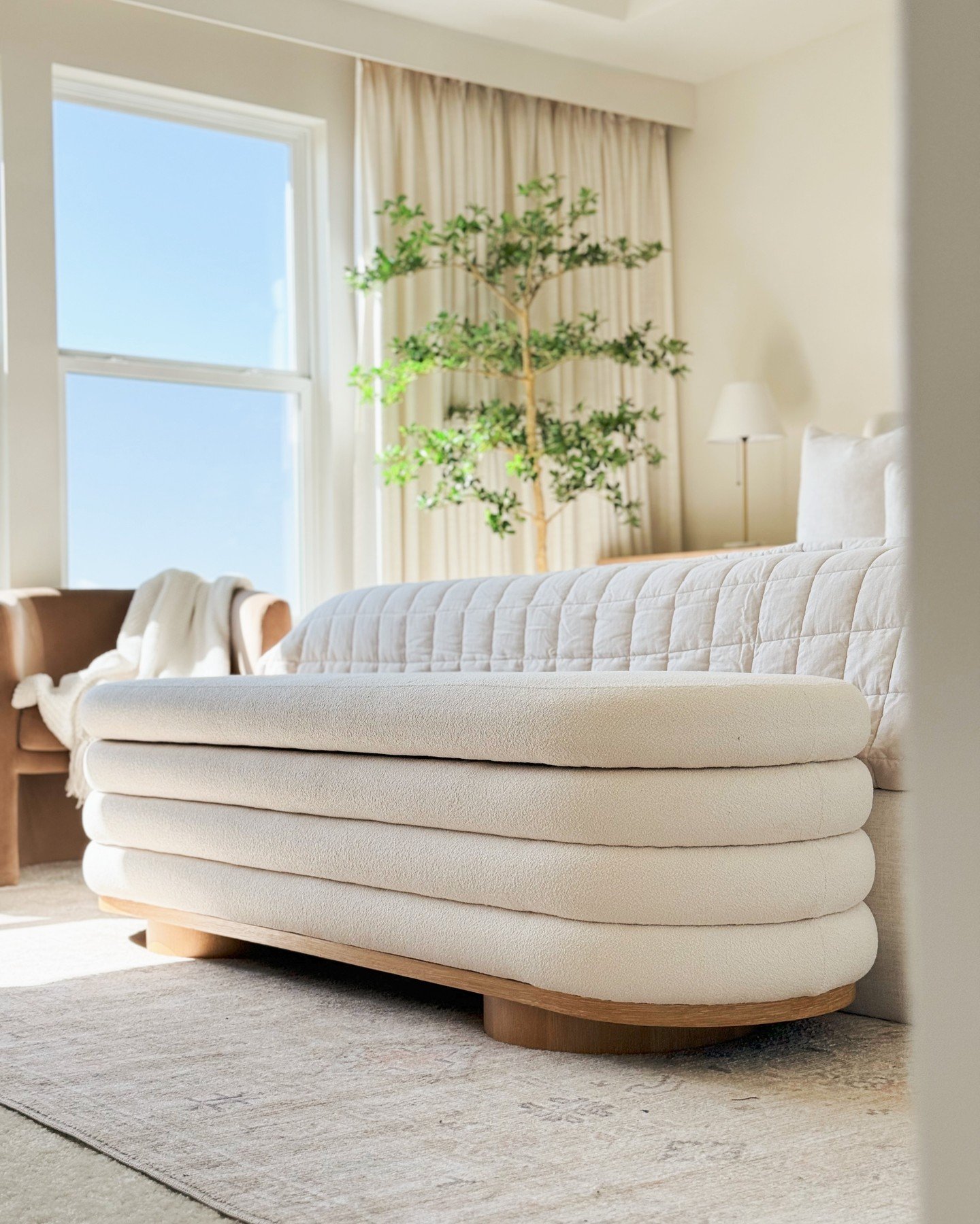 A close-up view of a modern, low-profile bedroom bench upholstered in thick, cream-colored bouclé fabric with a curved wooden base.