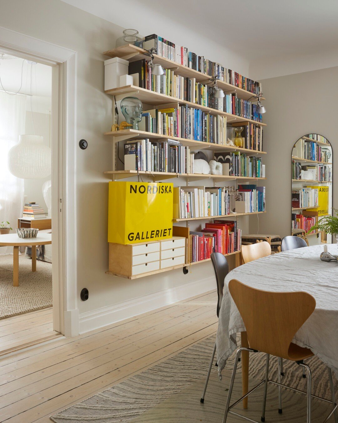 Modular shelving with natural wood shelves and white wire standards mounted on a light gray wall in a dining area, holding books, decor, and a large yellow storage bag.