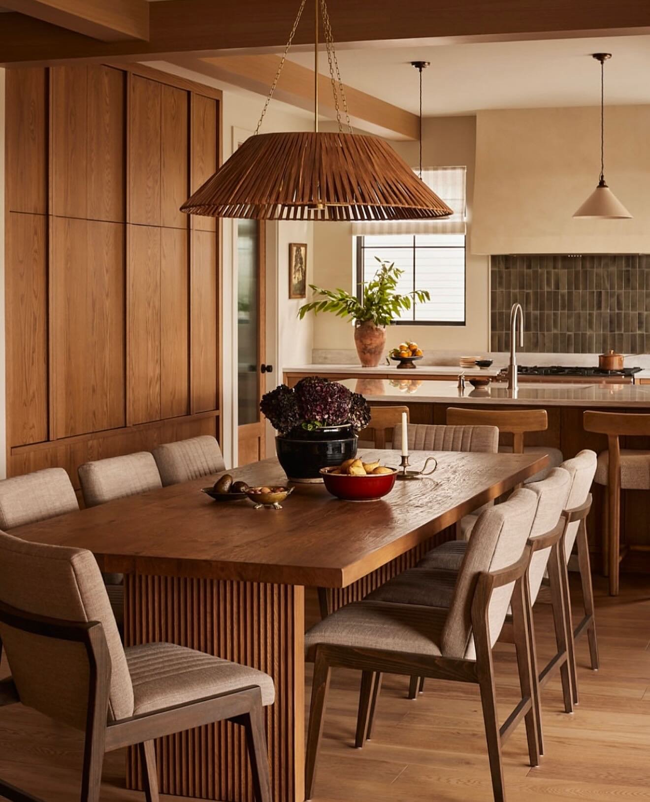A warm, open-plan dining area with floor-to-ceiling wood paneling, a large wooden table with a fluted base, and neutral upholstered chairs beneath a woven pendant light.