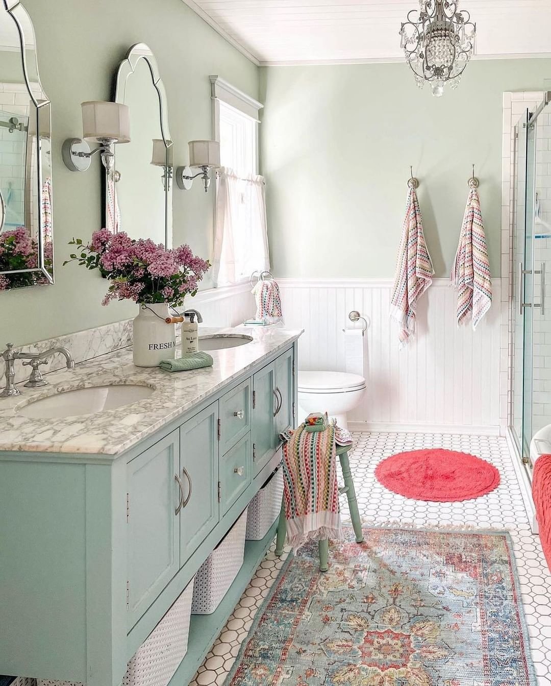 A mint green farmhouse bathroom featuring a marble-top vanity, white shiplap wainscoting, a hexagonal tile floor, and two layered rugs: a large Persian-style runner and a small, bright magenta circular rug.