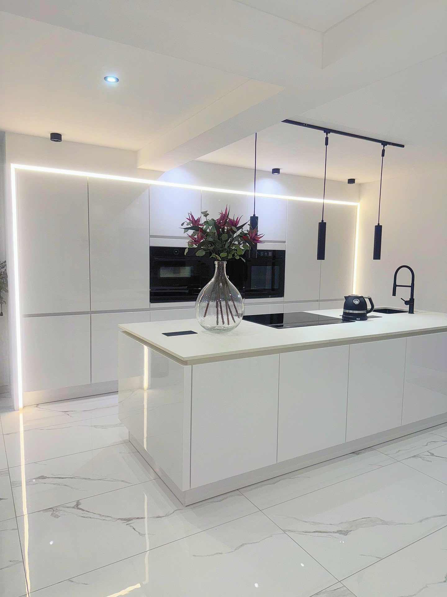 Ultra-modern kitchen featuring handleless white high-gloss cabinets, a large island with an integrated hob, black fixtures, and dramatic perimeter LED lighting.