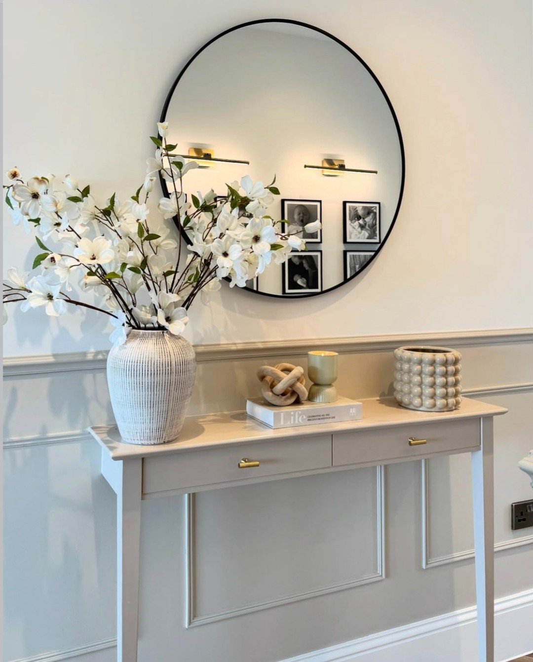 A narrow, light taupe console table with gold handles topped with white flowers a round black mirror and layered decorative items.